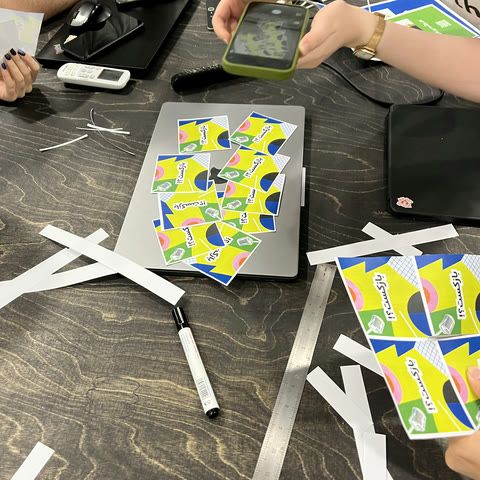 A desk mid-make: yellow and blue printed cards cut into pieces, a scalpel, a steel ruler and offcuts of paper
